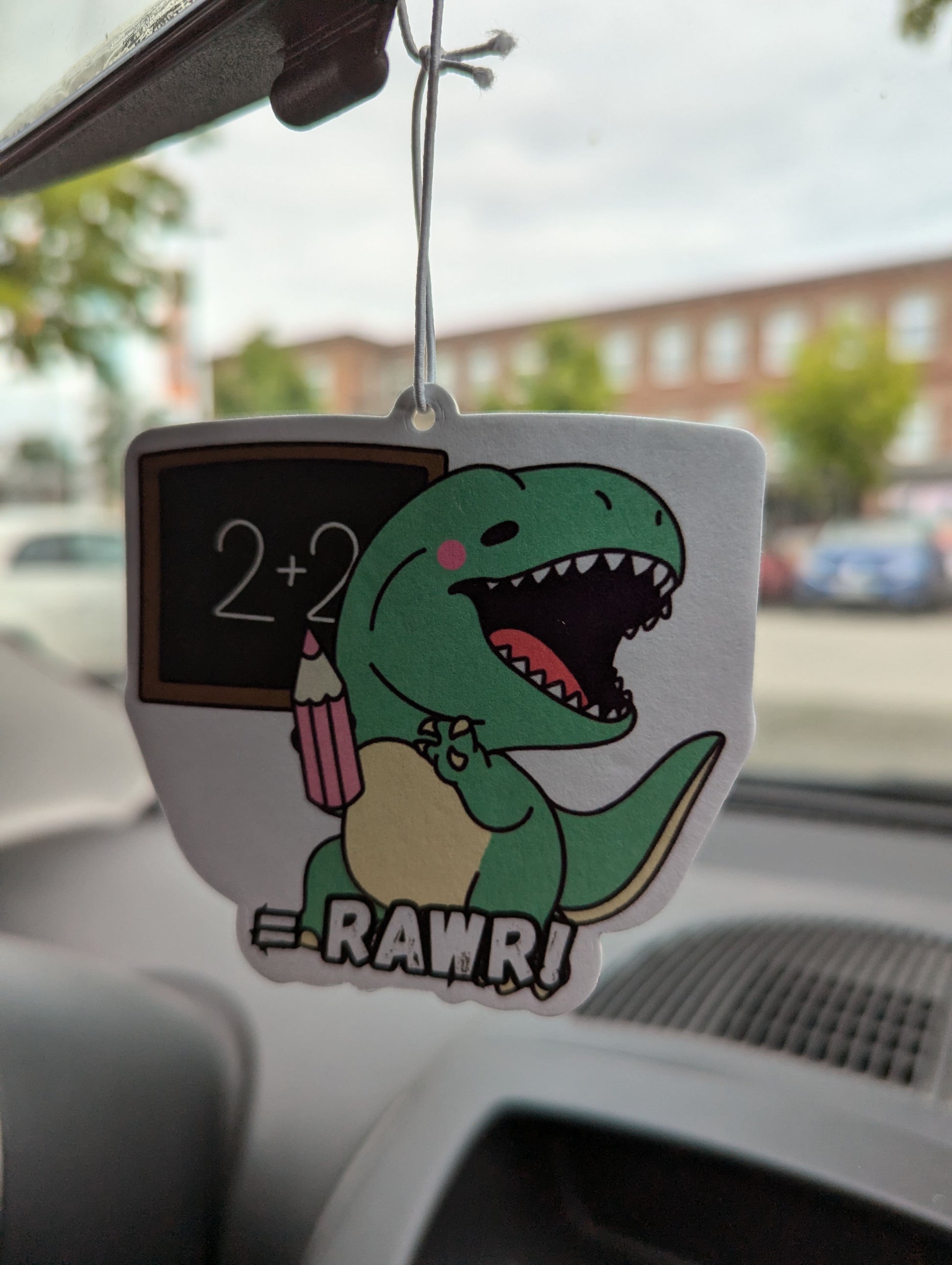 Auto Duftbaum T-Rex, zeigt auf eine Tafel und hängt im Auto am Rückspiegel