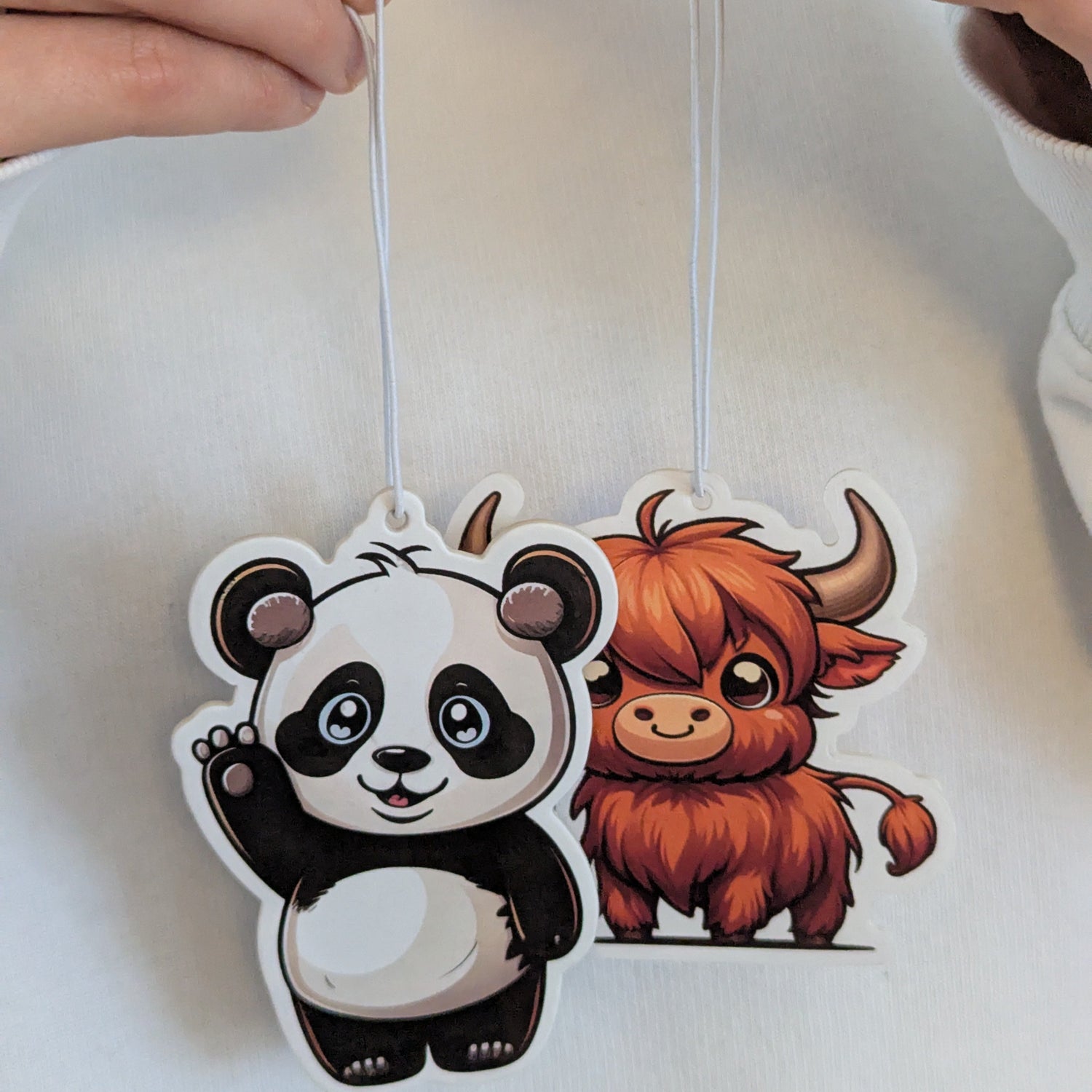 Zwei Auto Duftbäume nebeneinander – süßer Panda mit erhobener Pfote und freundliches Hochlandrind mit braunem Fell, Lufterfrischer fürs Auto mit Tiermotiven, dekorative Autoduftbäume als Geschenkidee.