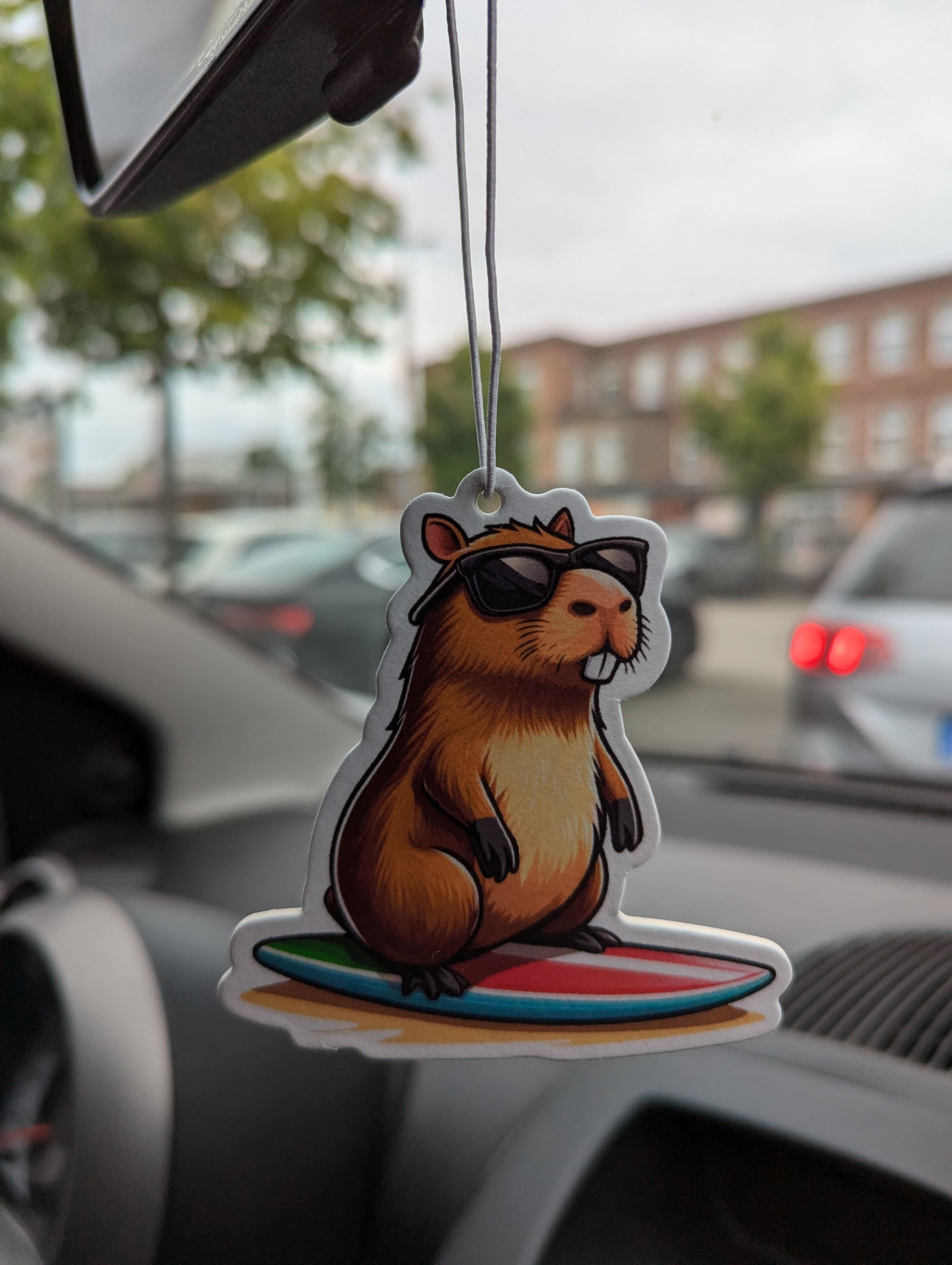 Auto Duftbaum Capybara mit schwarzer Sonnenbrille steht auf einem Surfbrett und hängt am Auto Rückspiegel
