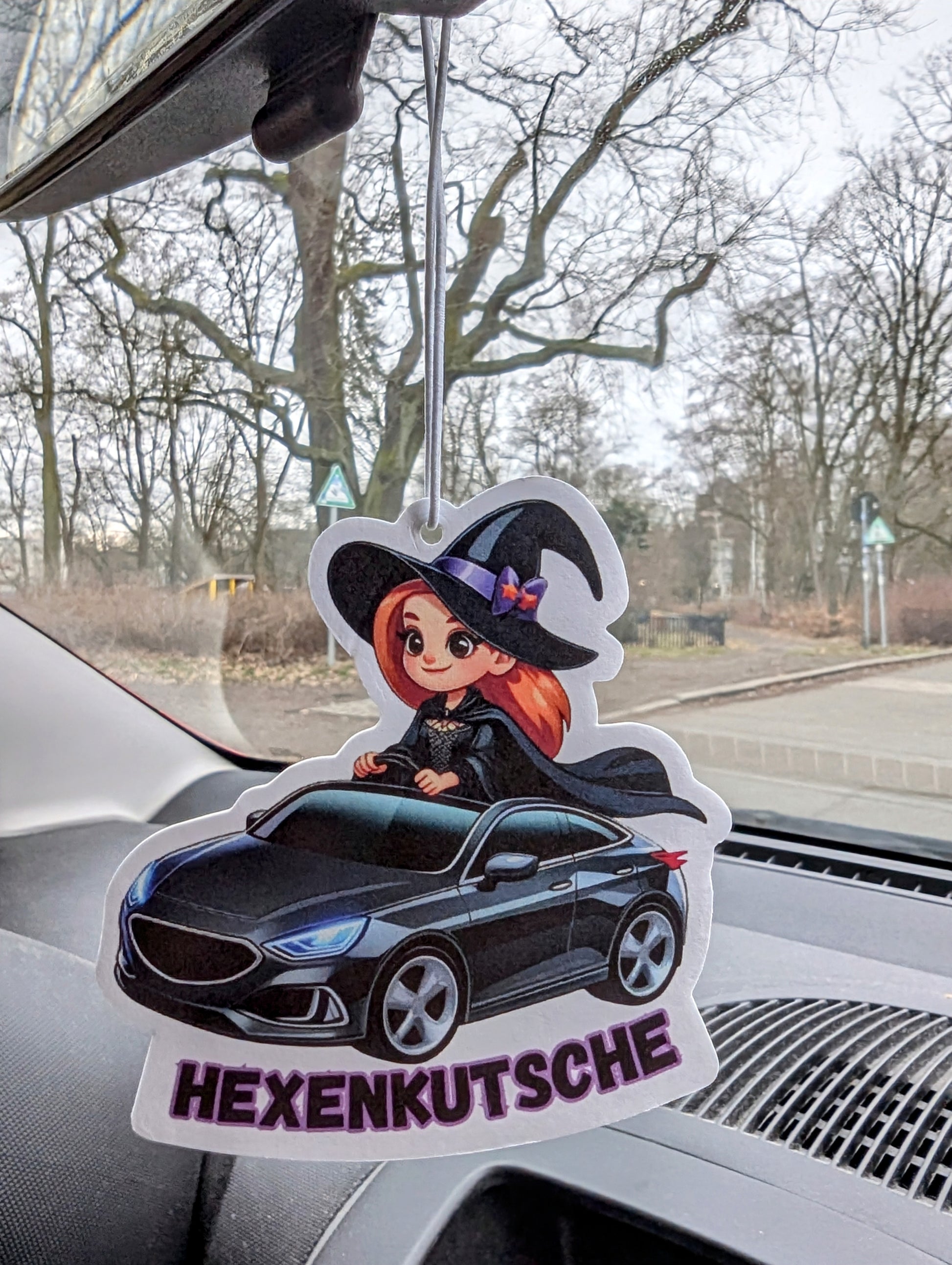Auto Duftbaum mit Comic-Hexe in schwarzem Kleid und spitzem Hut, die ein schwarzes Auto fährt, Schriftzug „Hexenkutsche“ darunter, Lufterfrischer fürs Auto mit witzigem Motiv als originelle Geschenkidee.