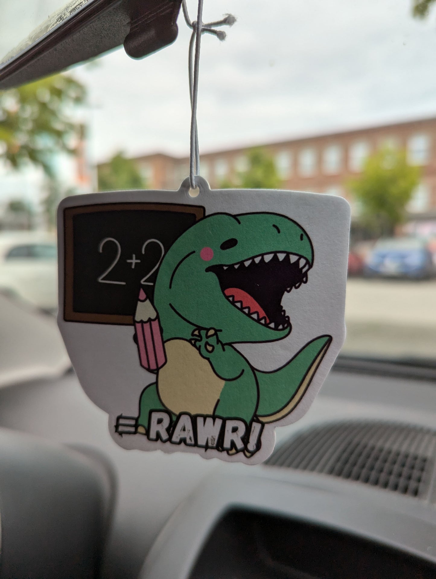 Auto Duftbaum T-Rex, zeigt auf eine Tafel und hängt im Auto am Rückspiegel